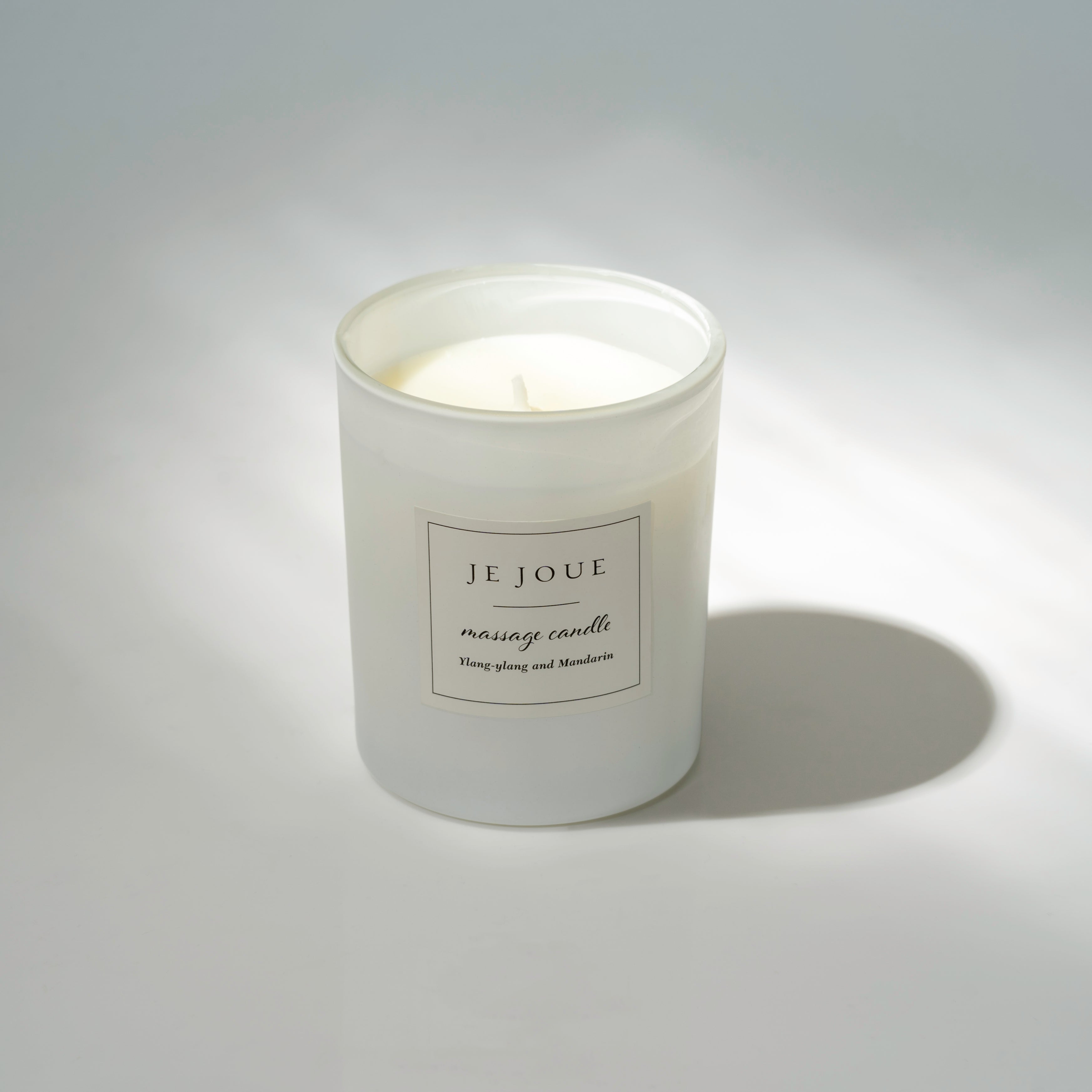 A white frosted glass massage candle with a label that reads "JE JOUE massage candle Ylang-ylang and Mandarin".
