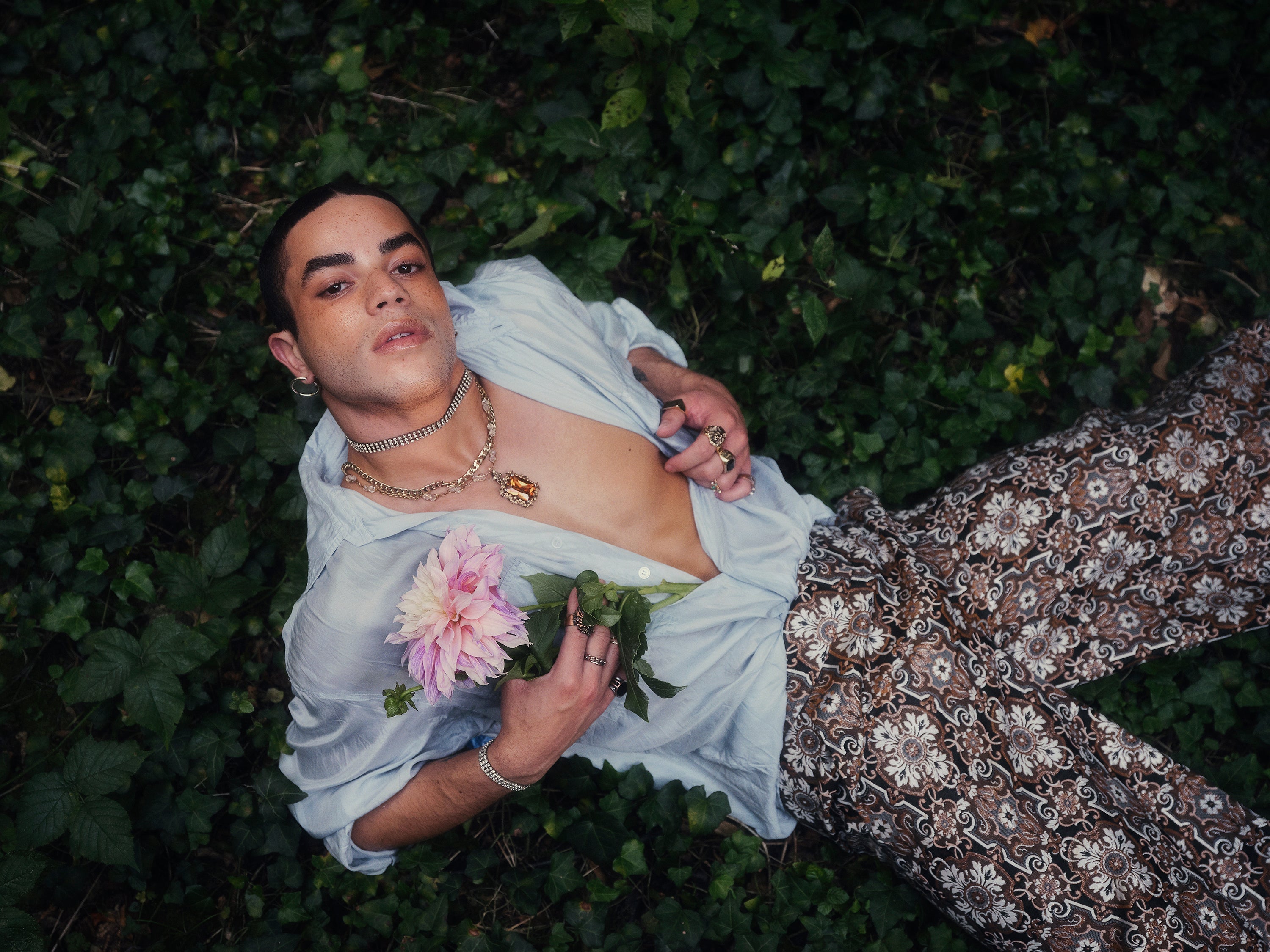 A person lies on green foliage wearing a light blue shirt and patterned pants, holding a pink flower.