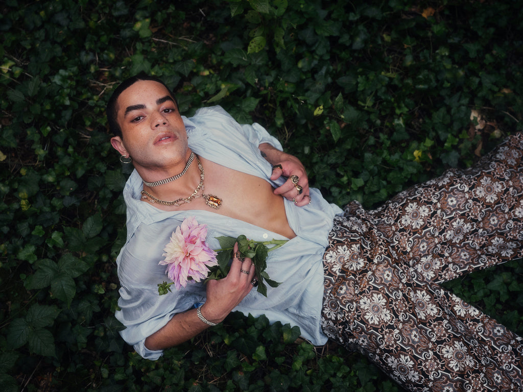A person lies on green foliage wearing a blue shirt and patterned pants, holding a pink flower.