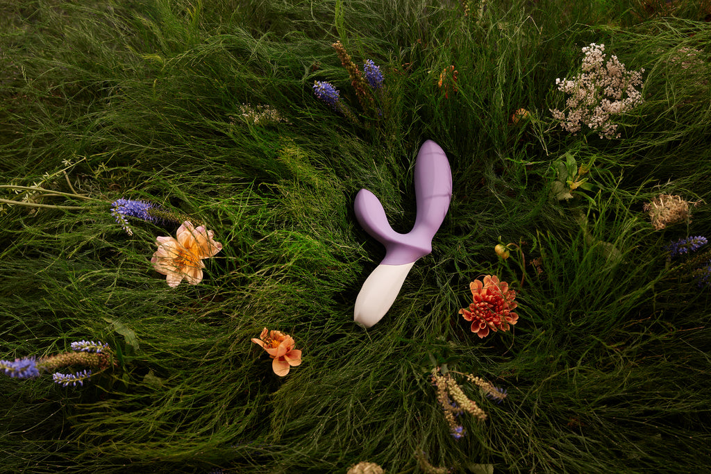 A purple and white prostate massager rests on a bed of green grass and flowers.