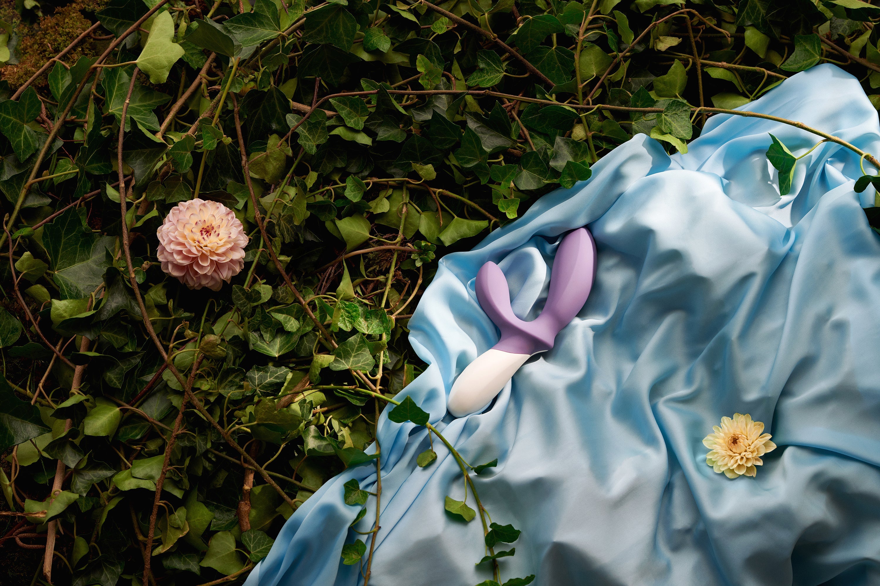A purple and white silicone prostate massager rests on a blue silk cloth amidst ivy and flowers.
