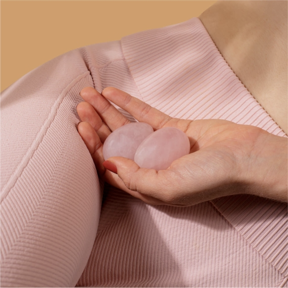 A hand holds two smooth, pink rose quartz yoni eggs.
