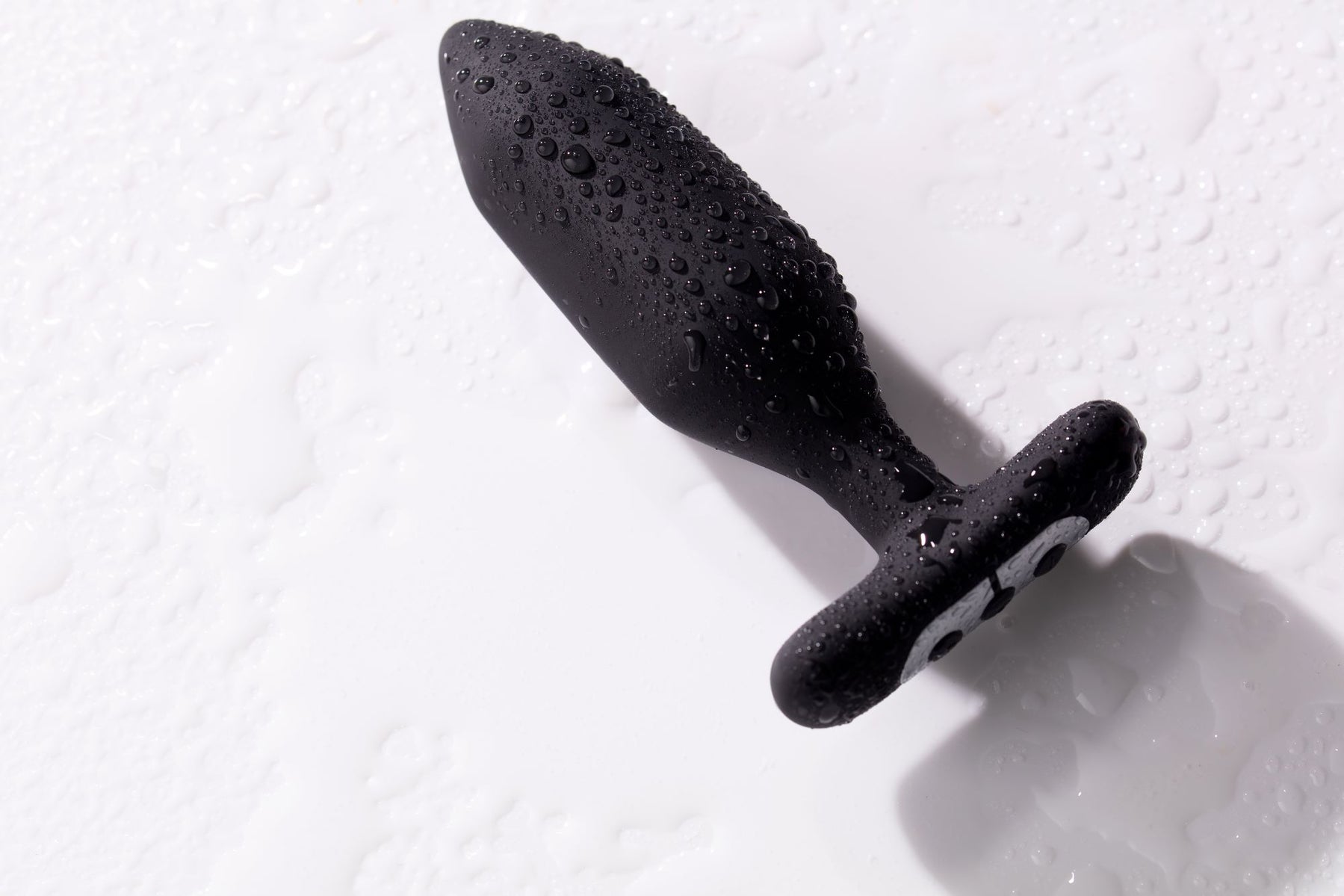 Black vibrating anal plug covered in water droplets on a white surface.
