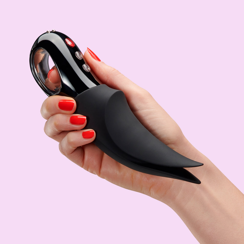 A hand holds a black vibrating pleasure set with a ring and curved handle.