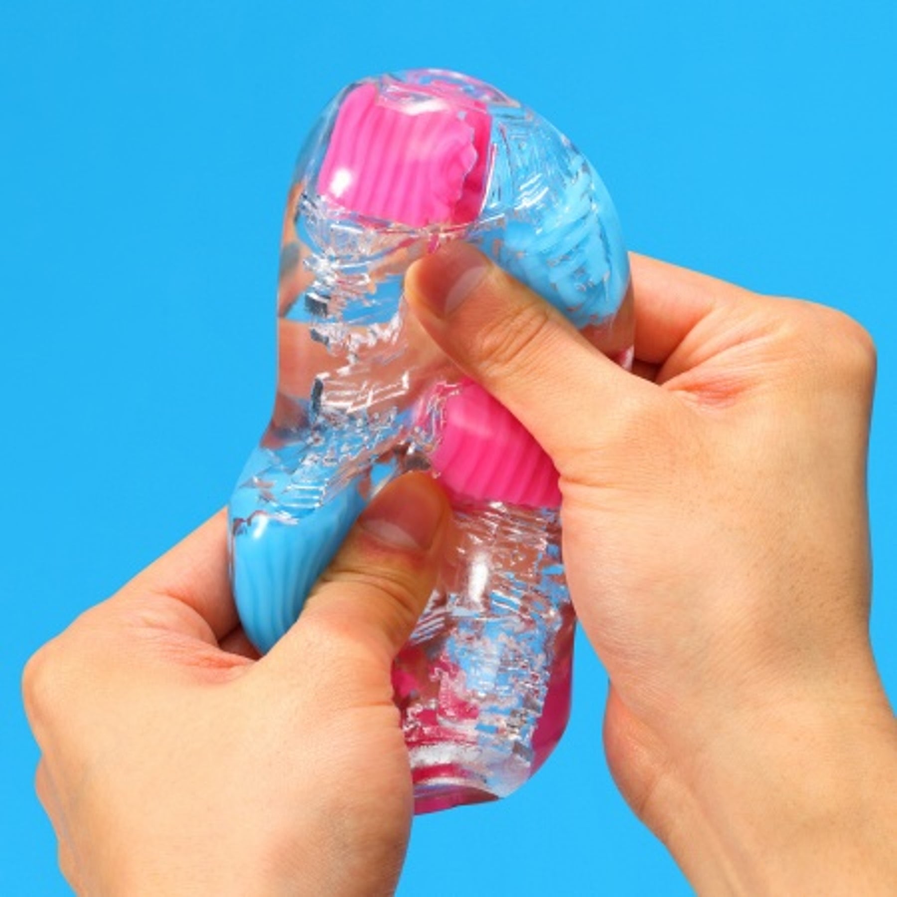 Hands squeezing a clear stroker with pink and blue textured inserts.