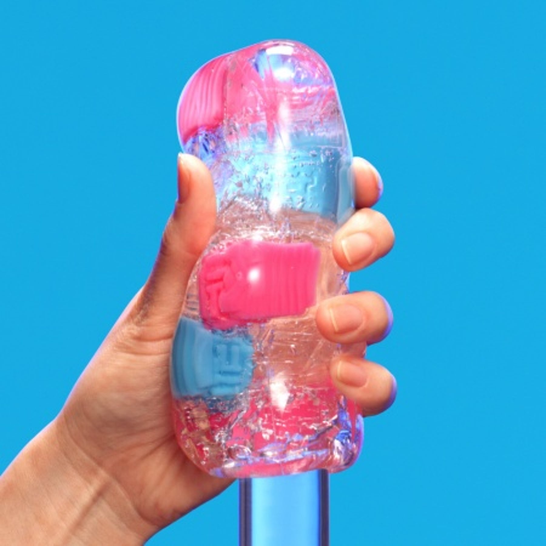 A hand holds a clear stroker with pink and blue textured inserts against a blue background.