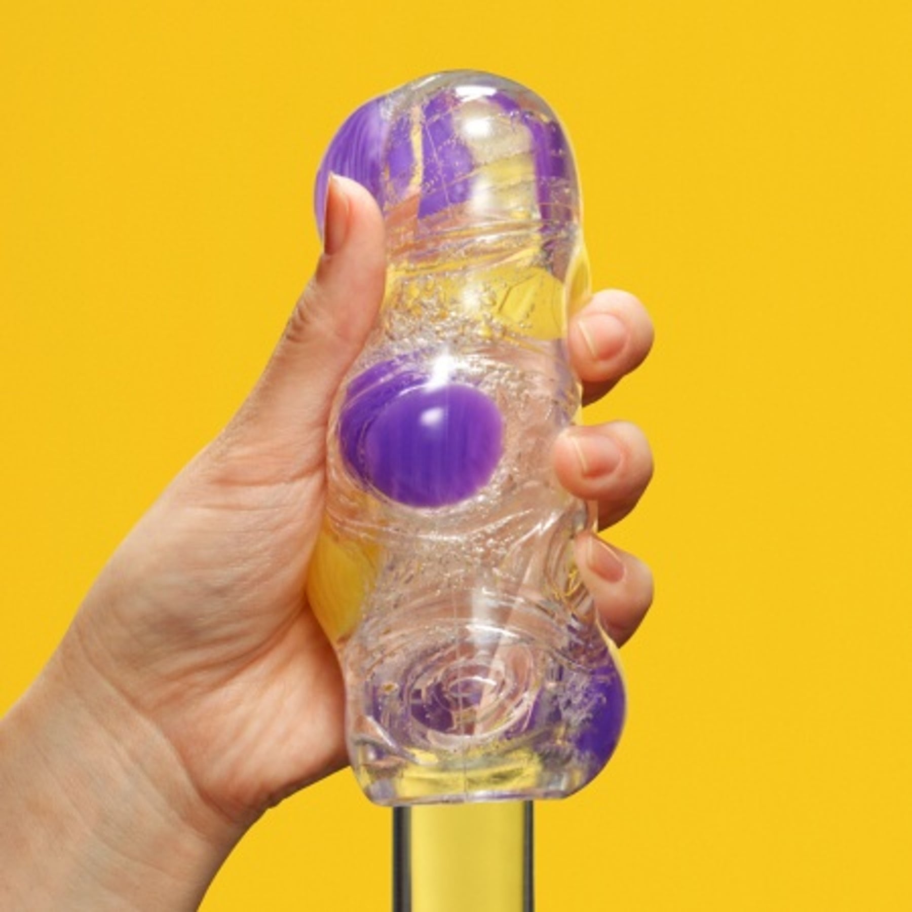 A hand holds a clear, textured stroker with purple marbles and liquid inside against a yellow background.