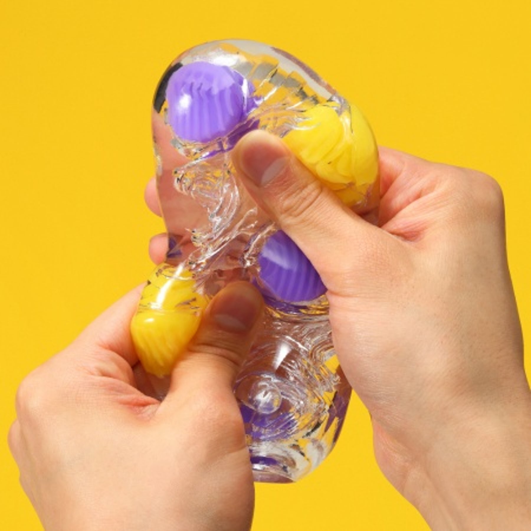 A clear textured stroker with purple and yellow bobbles is being squeezed by two hands.