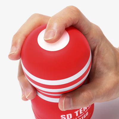 A hand grips a red Tenga masturbator with white stripes.