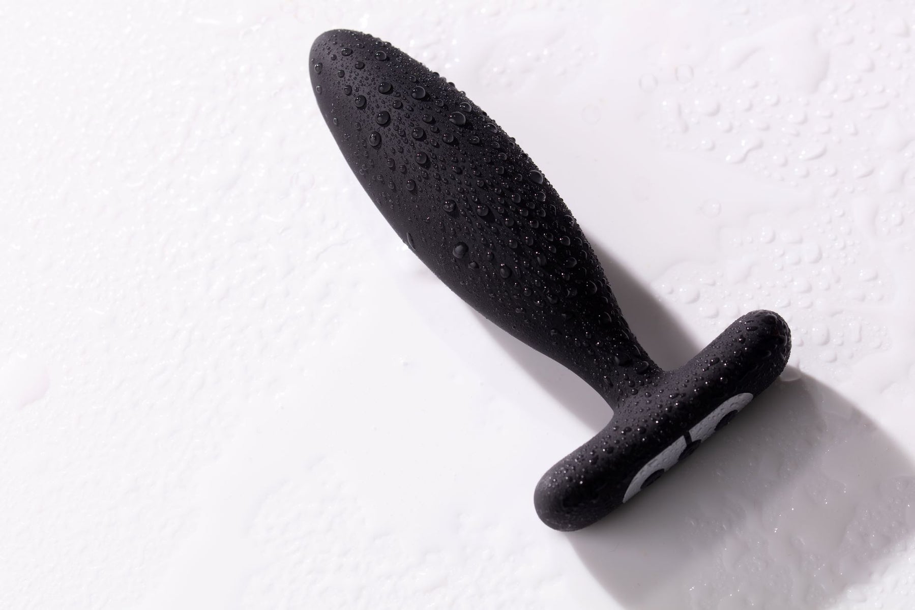 Black vibrating butt plug with water droplets on a white surface.