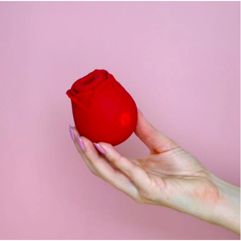 A hand holds a red rose-shaped massager against a pink background.