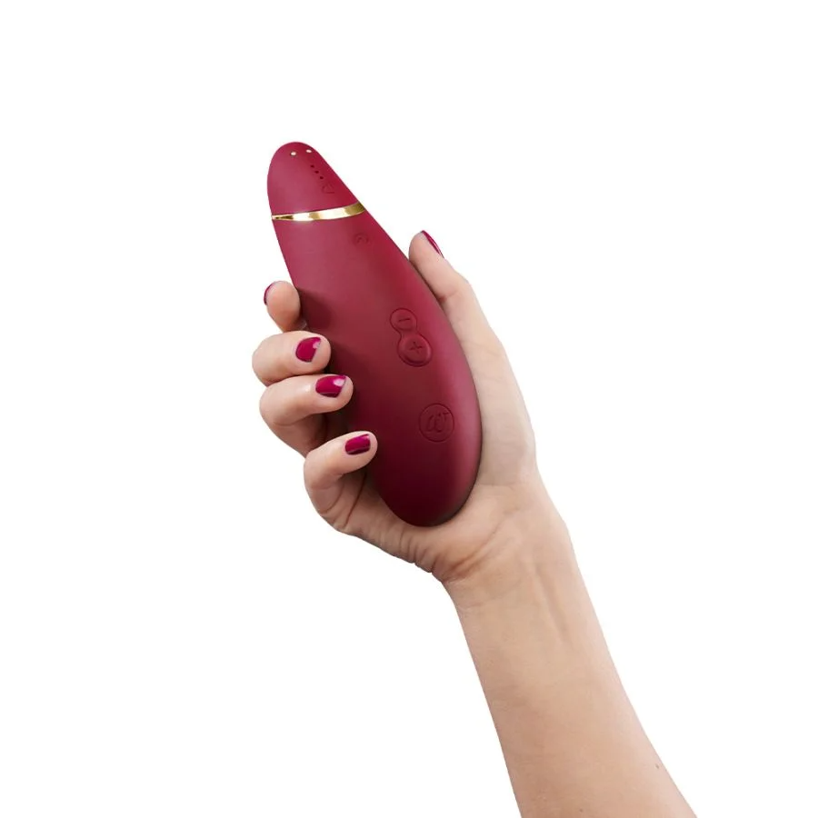 A hand holds a burgundy clitoral vibrator with gold accents and buttons.