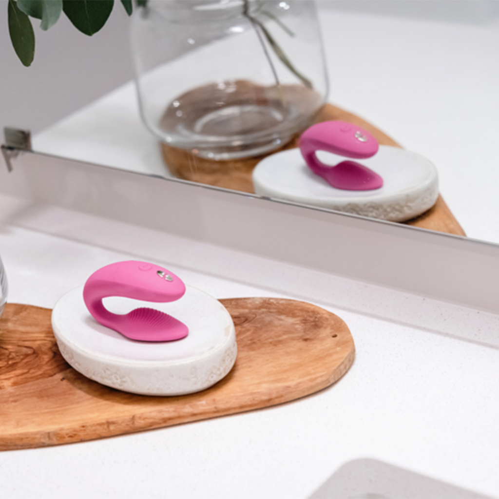 A pink app-controlled couples vibrator rests on a white stone.