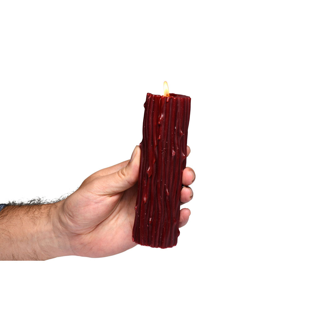 A hand holds a red pillar candle with a bark-like texture and a lit wick.
