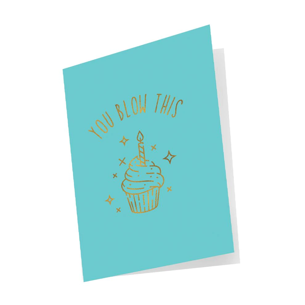 A turquoise greeting card with a gold foil illustration of a cupcake with a candle and the words "YOU BLOW THIS".